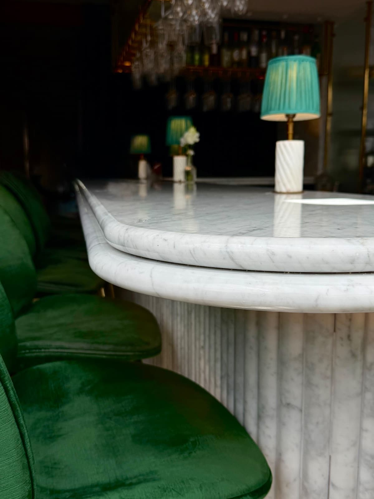 Close-up of a curved marble bar counter with emerald velvet stools and teal lamp shades