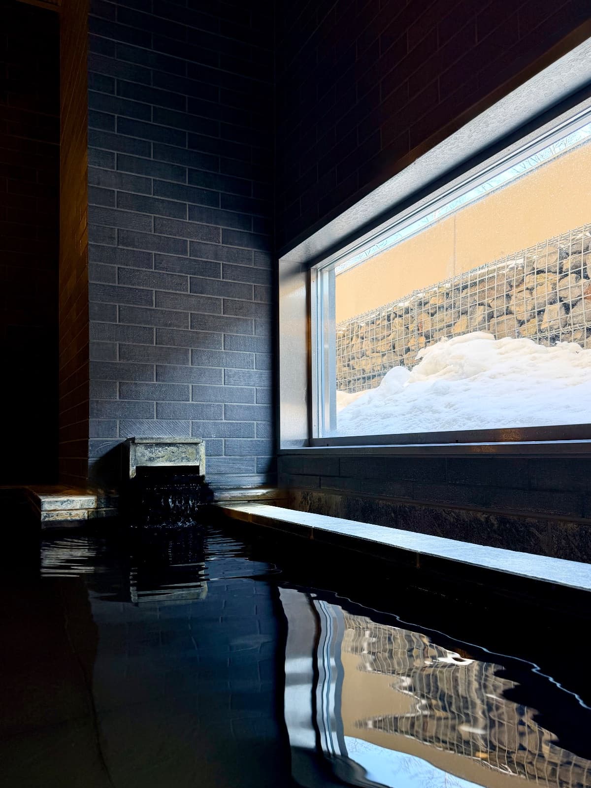 A dark stone onsen bath with still water reflecting a snowy window view in Japan
