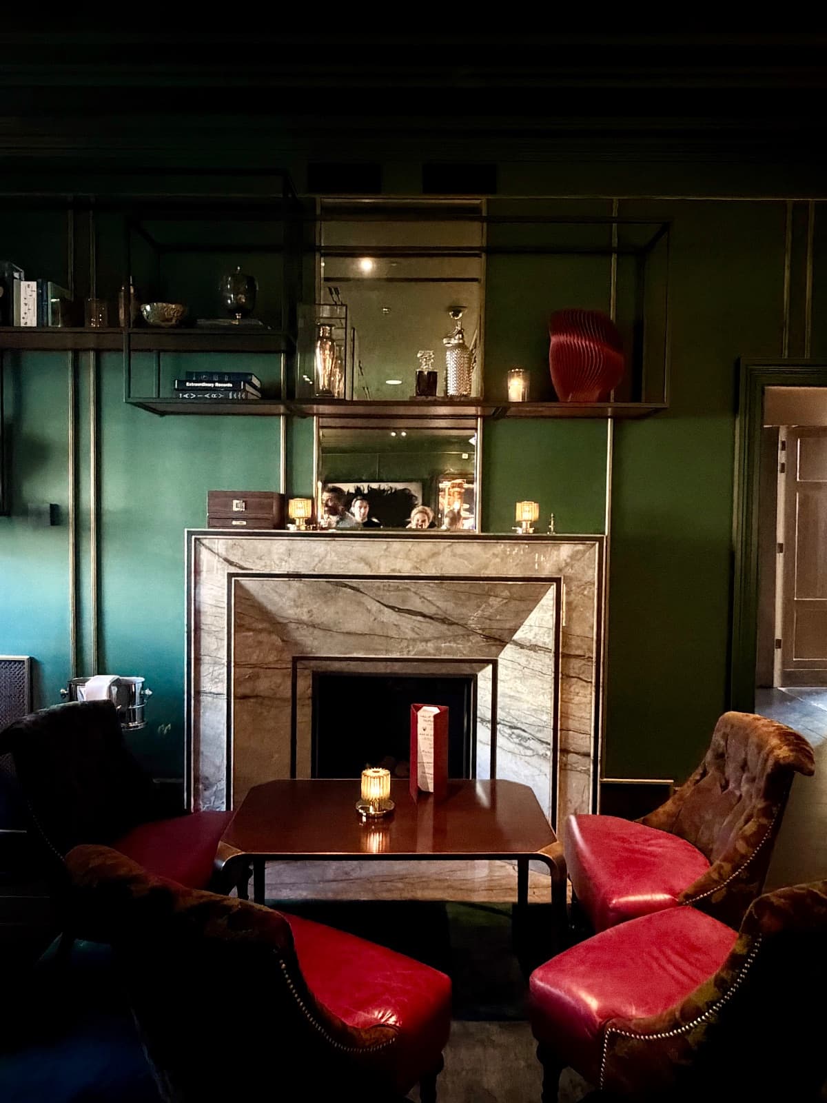 A moody green sitting room with a marble fireplace, red leather chairs, and brass shelving