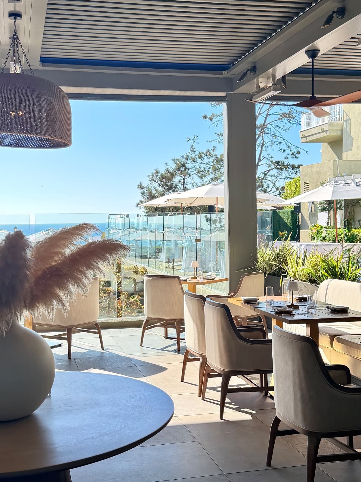 A sunlit oceanfront terrace with pampas grass, wood dining tables, and cream chairs overlooking the Pacific