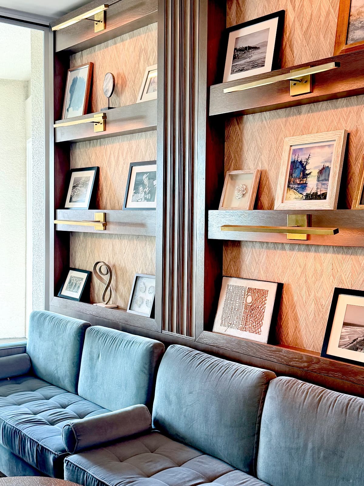 A curated wall of framed art and objects on wood shelving above a blue velvet sofa